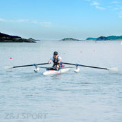 A person expertly navigates a scull boat using ZJ SPORTs ZJ Para-Rowing Oars with Carbon Oval Shaft on calm water, surrounded by small islands. The clear blue sky enhances the serene scene as the rower, in a cap and athletic wear, glides past various buoys.