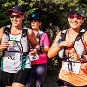Three women participate in an outdoor run wearing Z&J SPORT Unisex Quick Dry Adjustable Unstructured Caps by ZJ SPORT, smiling as they jog on a tree-lined path. Two runners wear white and orange tops, enjoying UV protection from their caps.