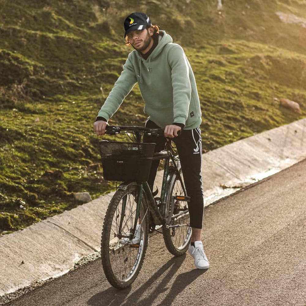 A person in a green hoodie, ZJ SPORT Unisex Quick Dry Adjustable Cap for outdoor sports, and glasses stands beside a bicycle on a paved road. They wear black pants and white sneakers, with green grass and sunlit hills in the background.
