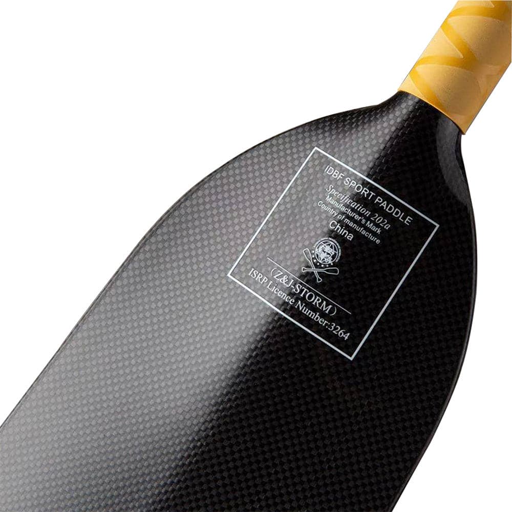 Close-up of the ZJ SPORT STORM paddle with a black carbon fiber dihedral blade featuring a textured pattern and labeled specs. The handle is wrapped in yellow tape for an anti-skid grip, highlighting its IDBF-approved quality for top-tier performance.